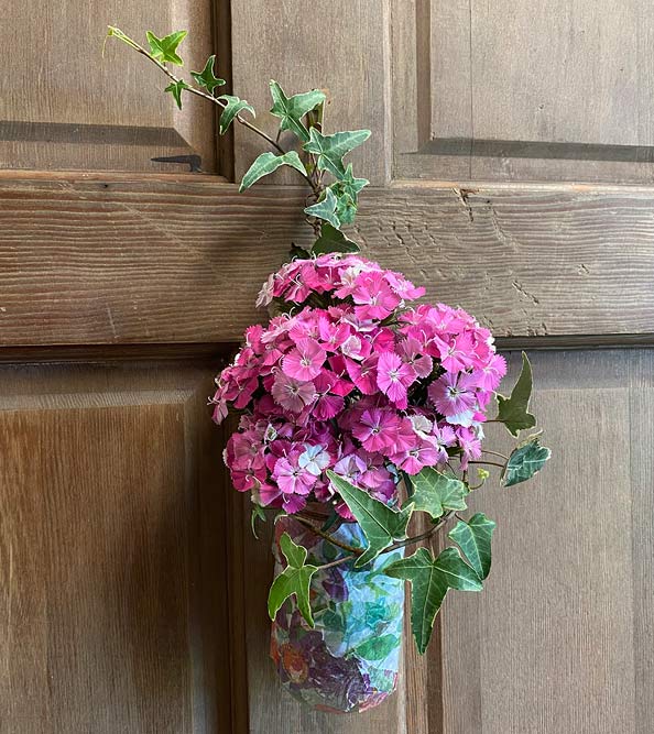 Small arrangement of pink dianthus blooms and ivy designed within glass jar hung on door.
