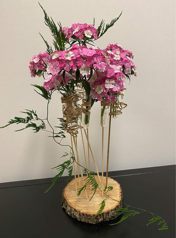 Dianthus centerpiece arrangement featuring a tree trunk base, green decorative foliage and pink blooms attached to wooden stems.
