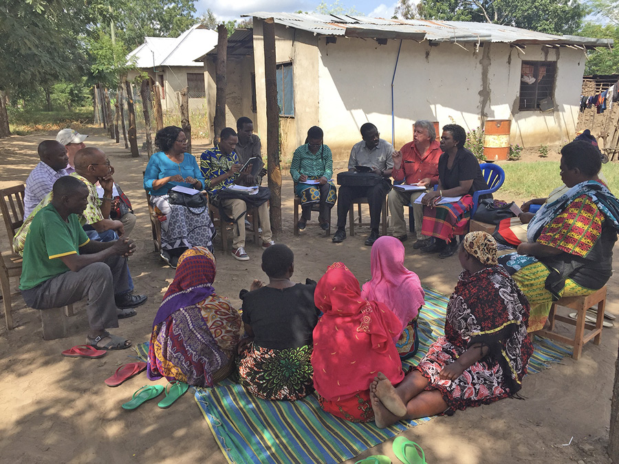 A group of people sit in a circle outside, some in chairs and some on the dirt ground. One person is talking and several others are taking notes.