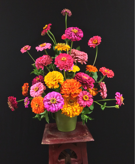 An arrangement of many yellow, pink, and orange zinnias in a green vase on a wood plant stand.
