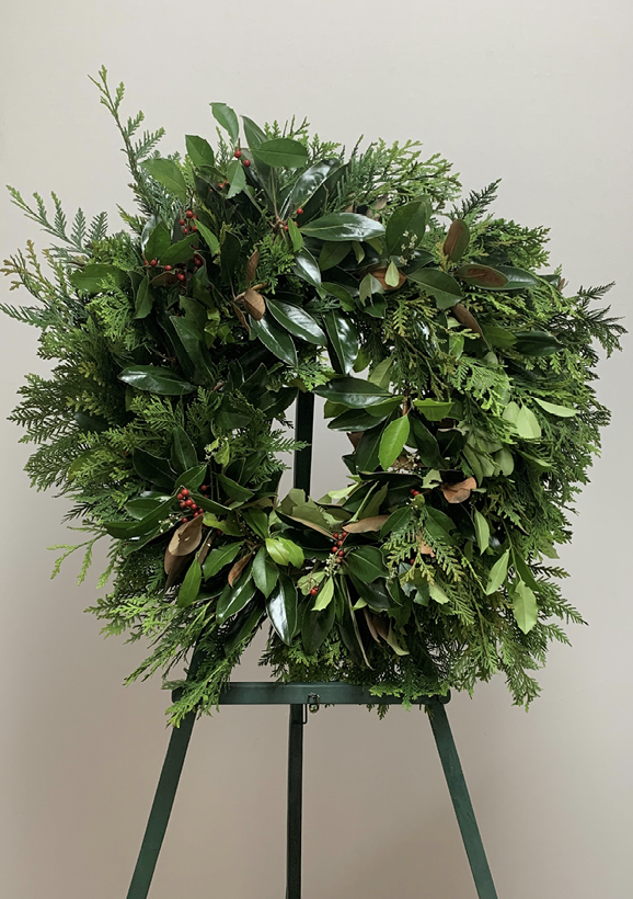 A wreath of magnolia leaves and other greenery and red berries displayed on a wreath stand.
