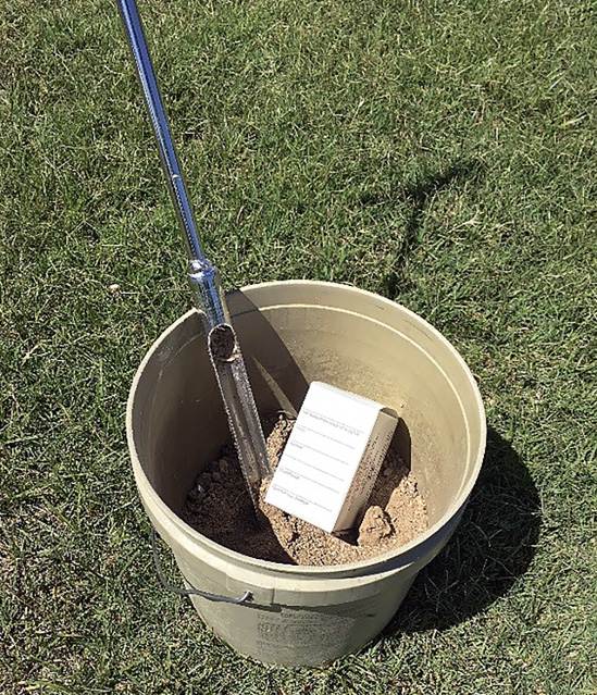 A bucket with soil, a soil probe, and a soil sample box.