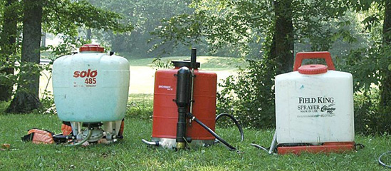Three different backpack sprayers displayed in a pecan orchard.