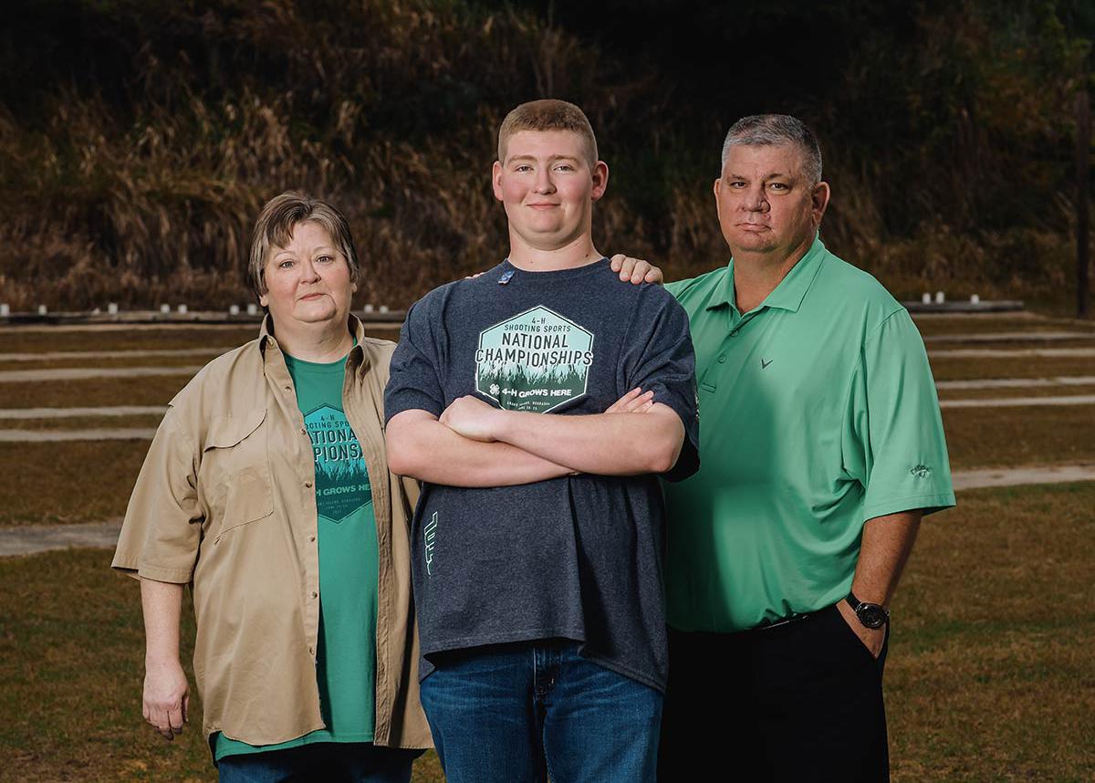 A teenage boy wearing a Shooting Sports National Championships shirt stands between his mother and father as they all smile.