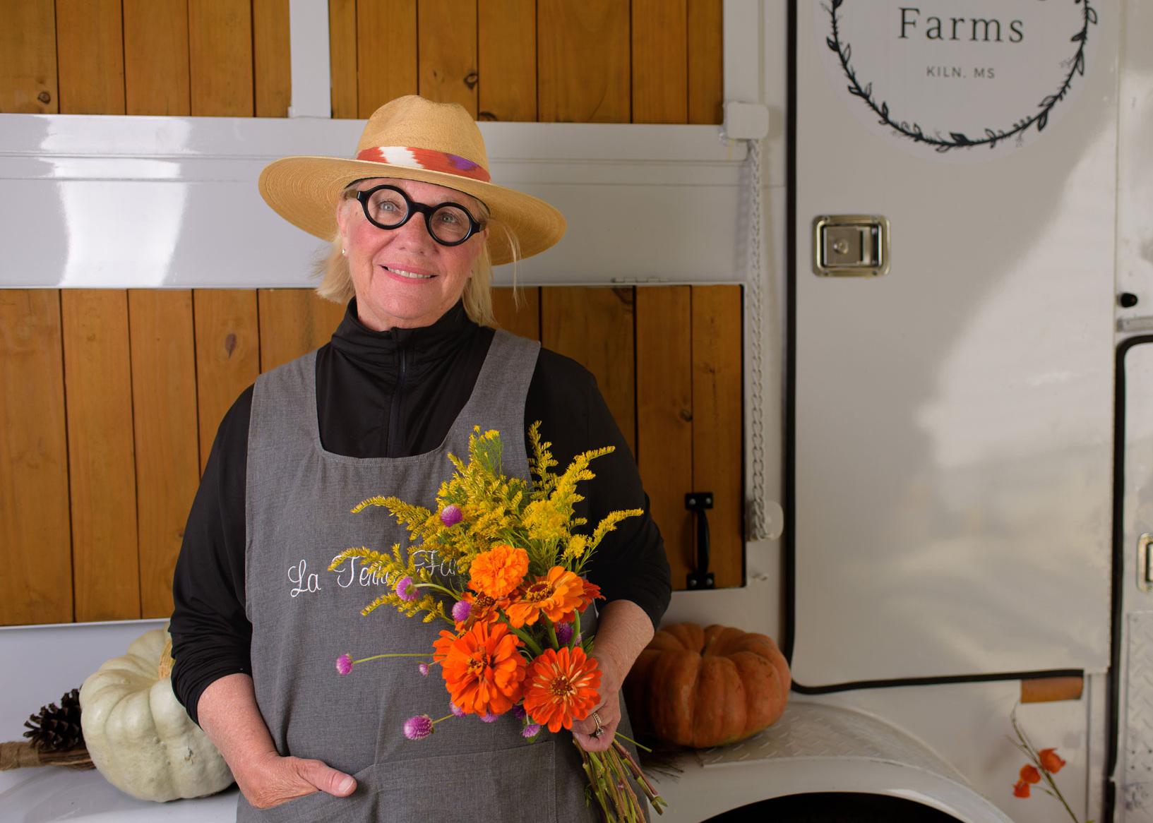 A woman with a straw hat and round-framed glasses holding a bunch of flowers and smiling.