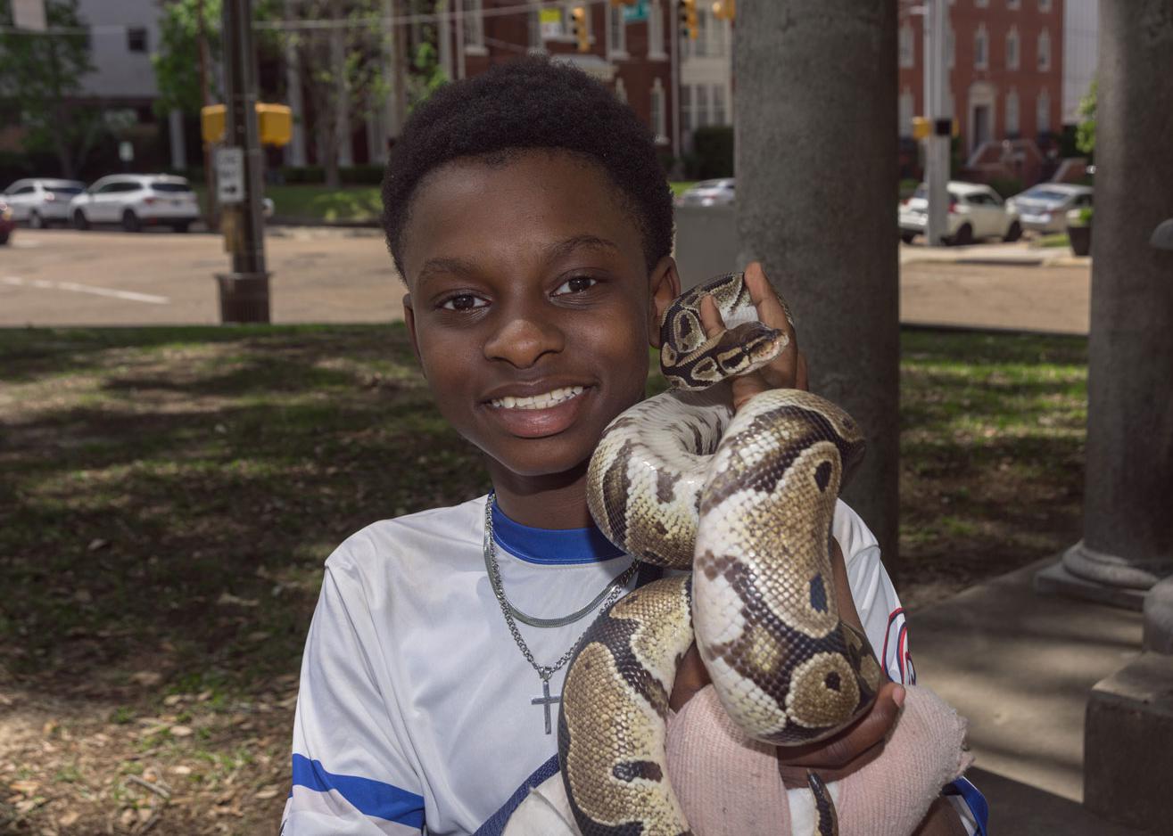A young smiling Black man holding a snake and standing near a city street.