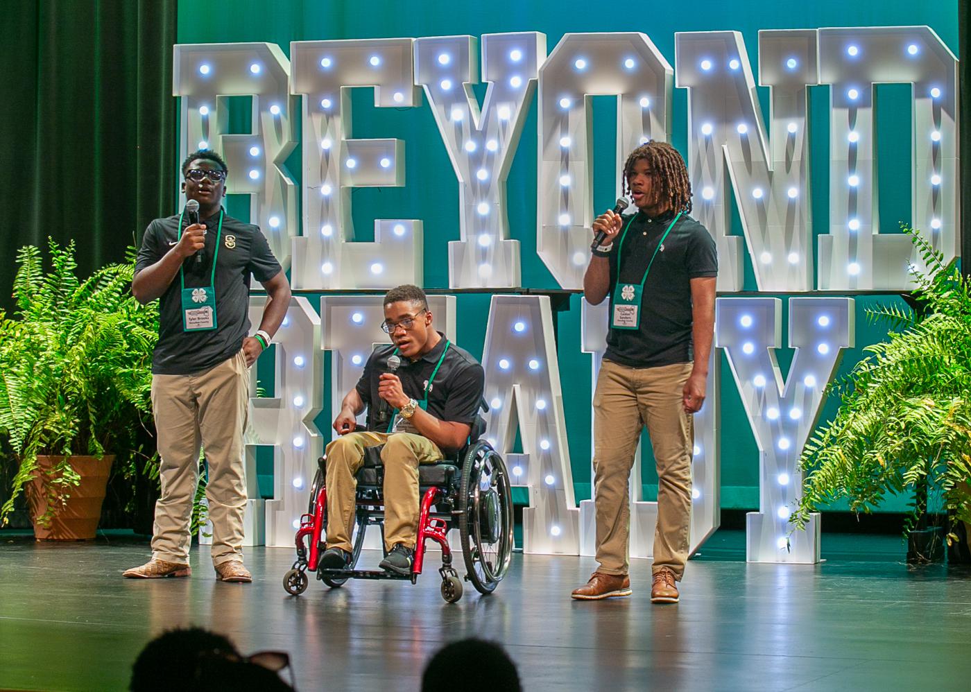 Three teens sing on stage, with the central performer seated in a wheelchair. Large, illuminated letters, arranged to form the words “Beyond Ready,” are in the background.