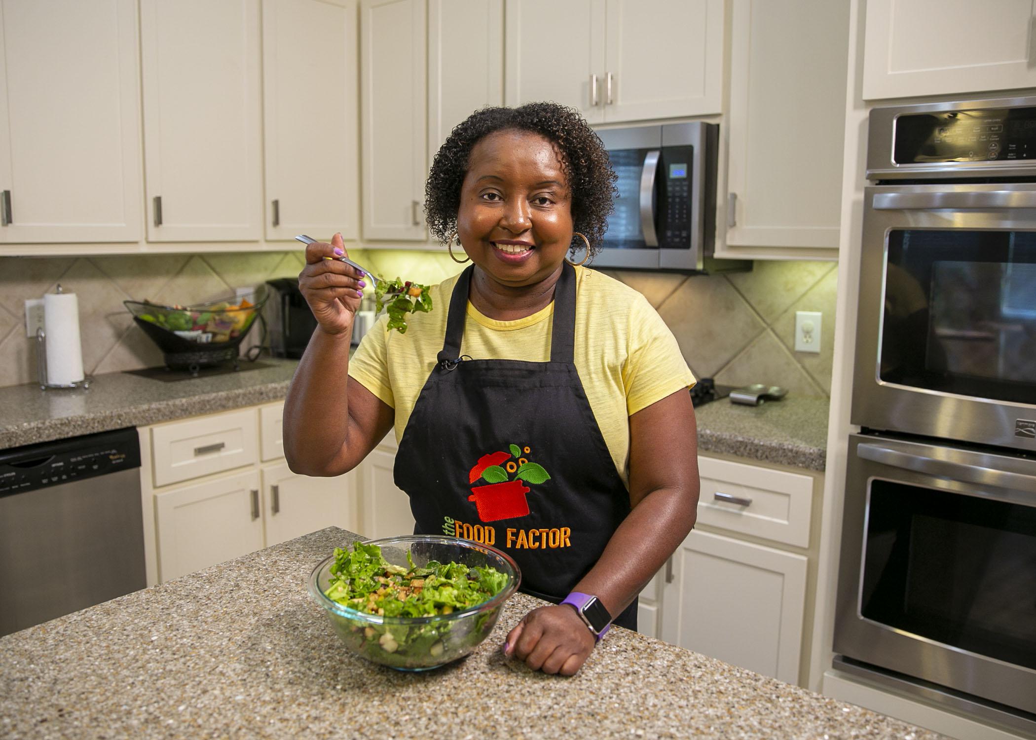 A woman in a kitchen smiles and holds a forkful of salad up for the camera.