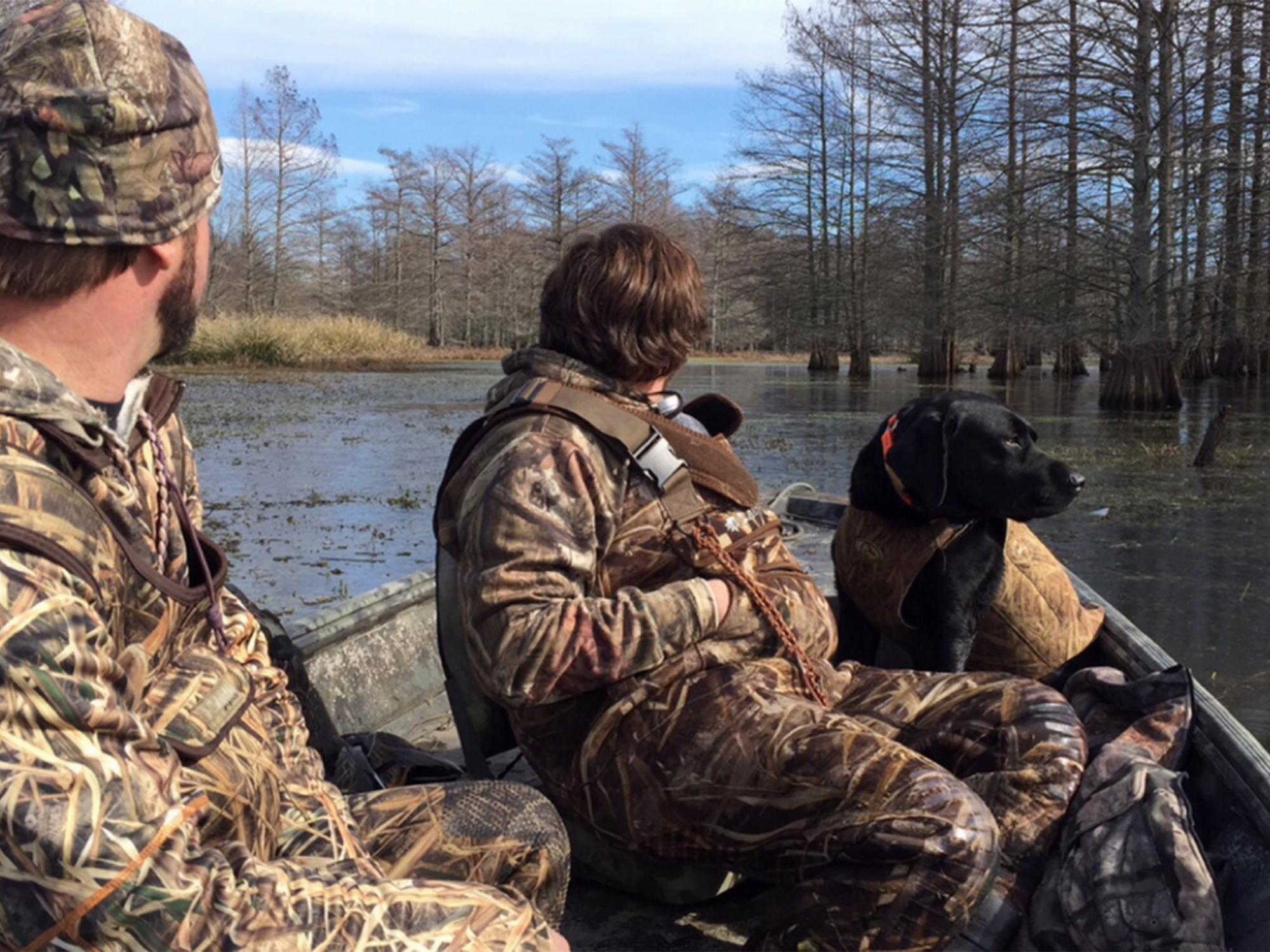 Two young men wearing camouflage sit in a small boat with a black dog, all looking out on the water on a sunny day.  