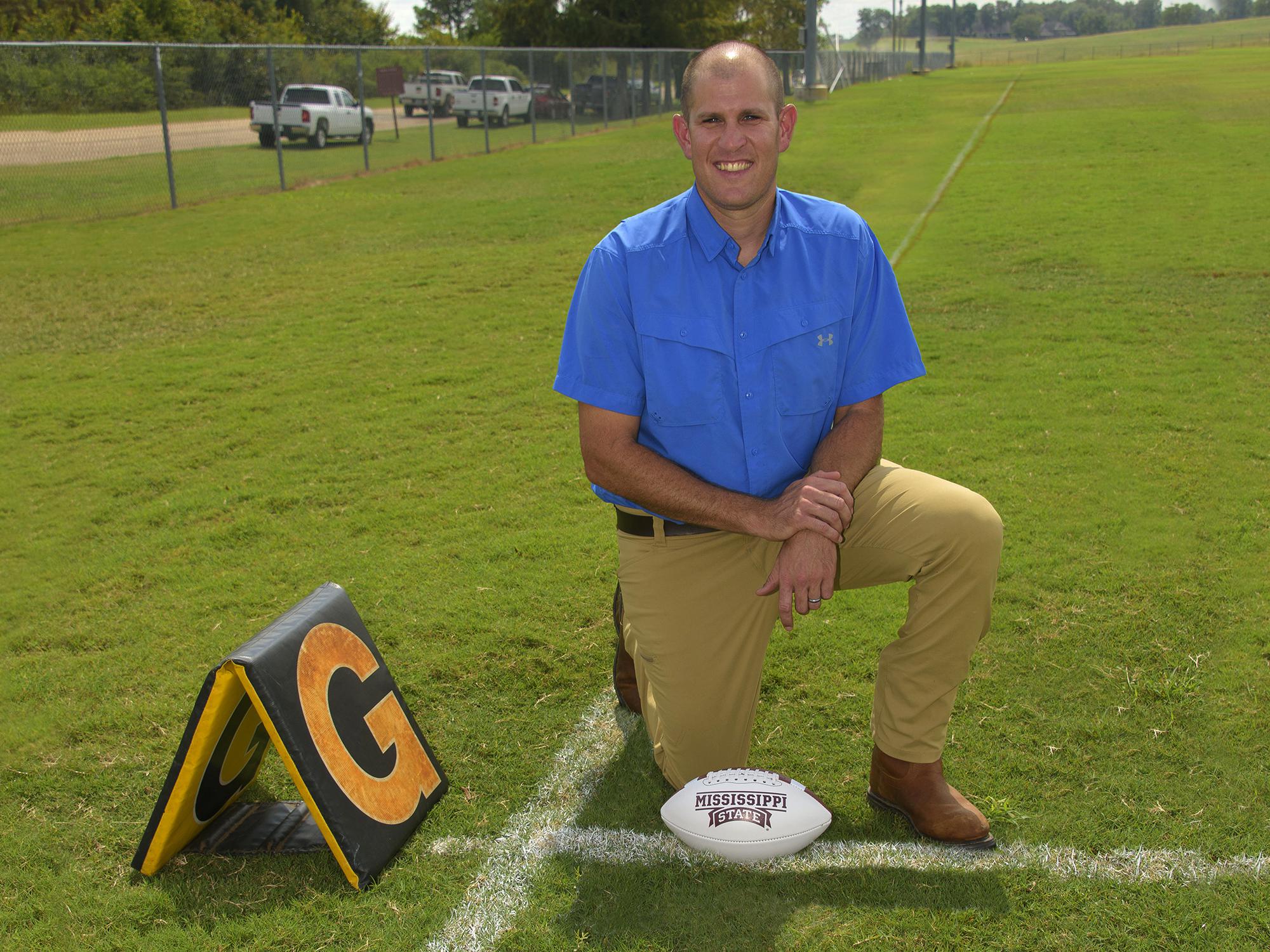 A man in a blue shirt kneels on a chalk-lined football field beside a goal line marker. A white Mississippi State football rests on the ground beside his knee.