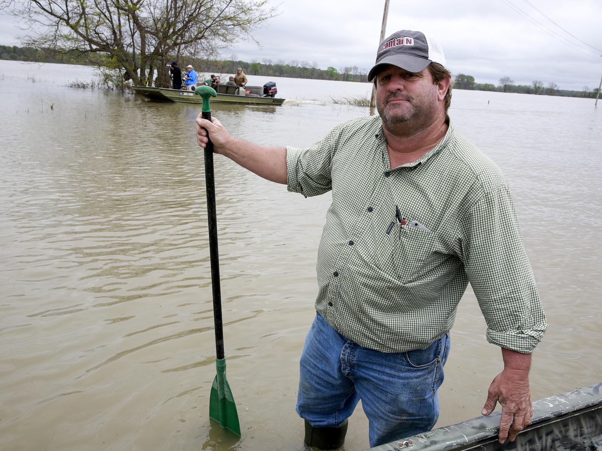 A man in work clothes, baseball cap and wading boots stands in water outside his boat holding a paddle.