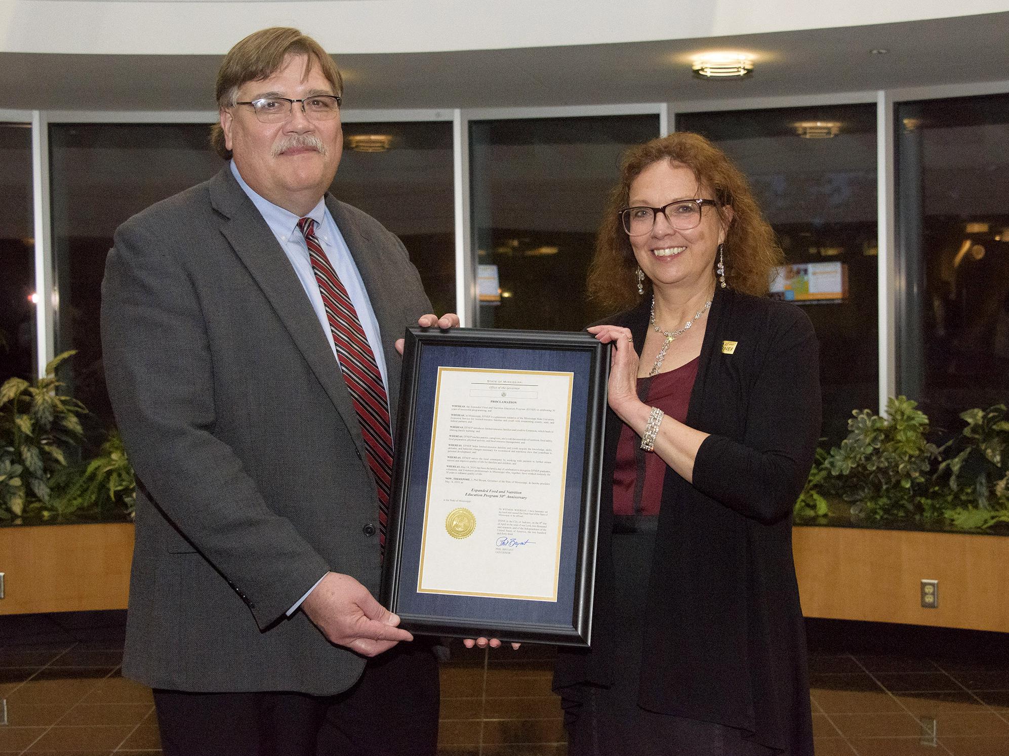 A man on the left and a woman on the right stand in an atrium and hold a framed document with a gold seal.