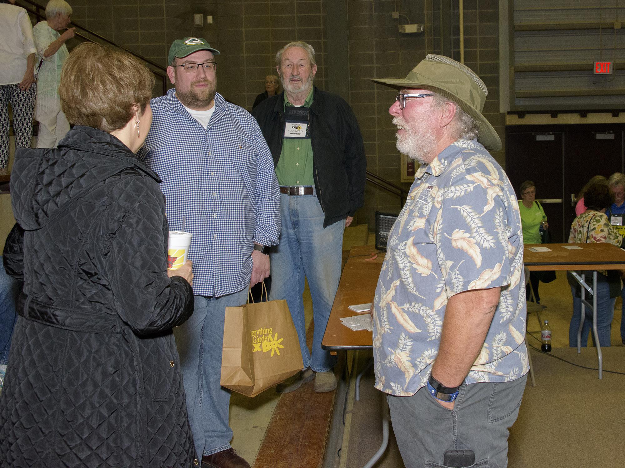 A man in a gardening hat stands and talks to three adults, with several others milling about in the background.