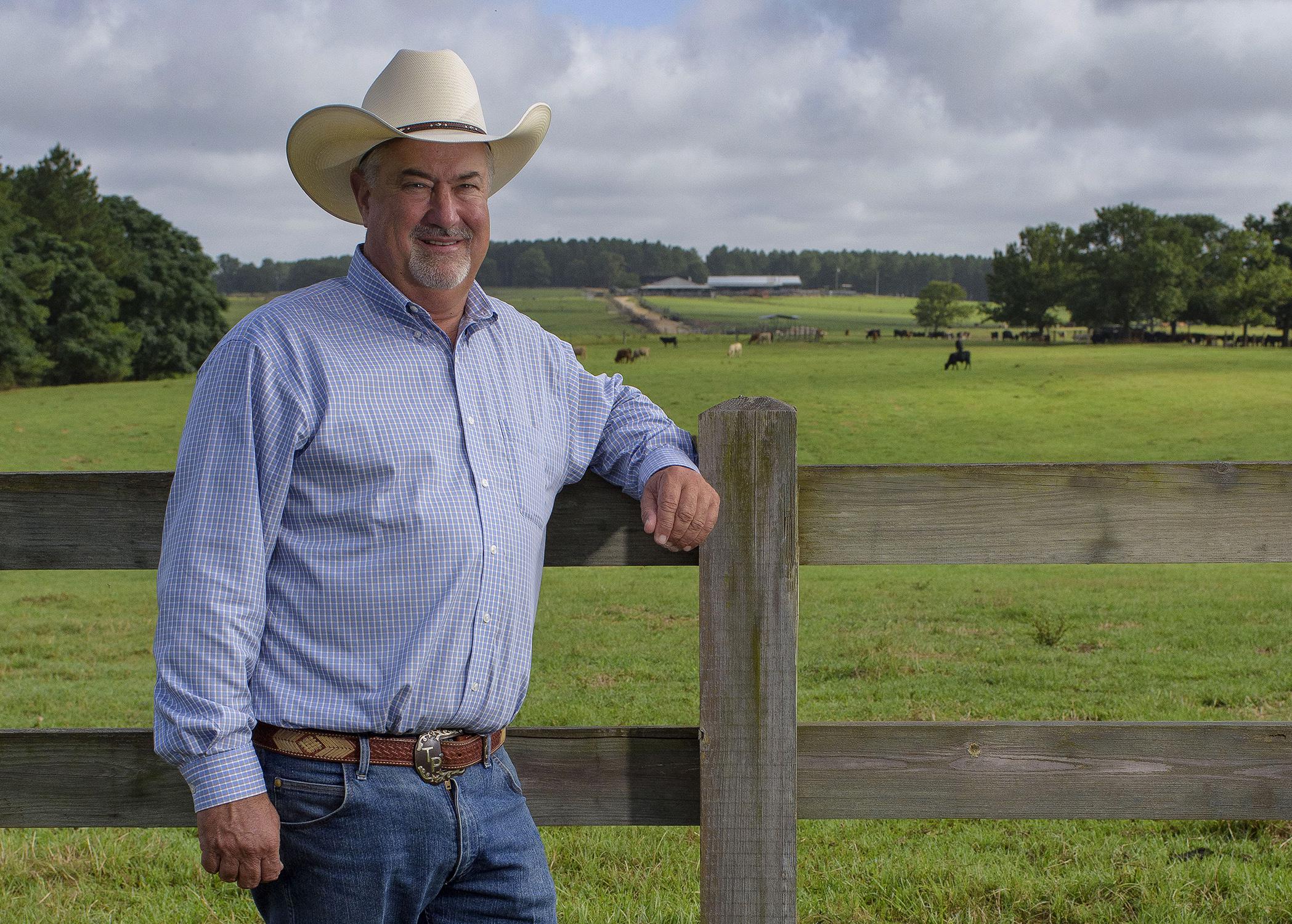 Man wearing a cowboy hat standing against a fence in front of a field with cows grazing.