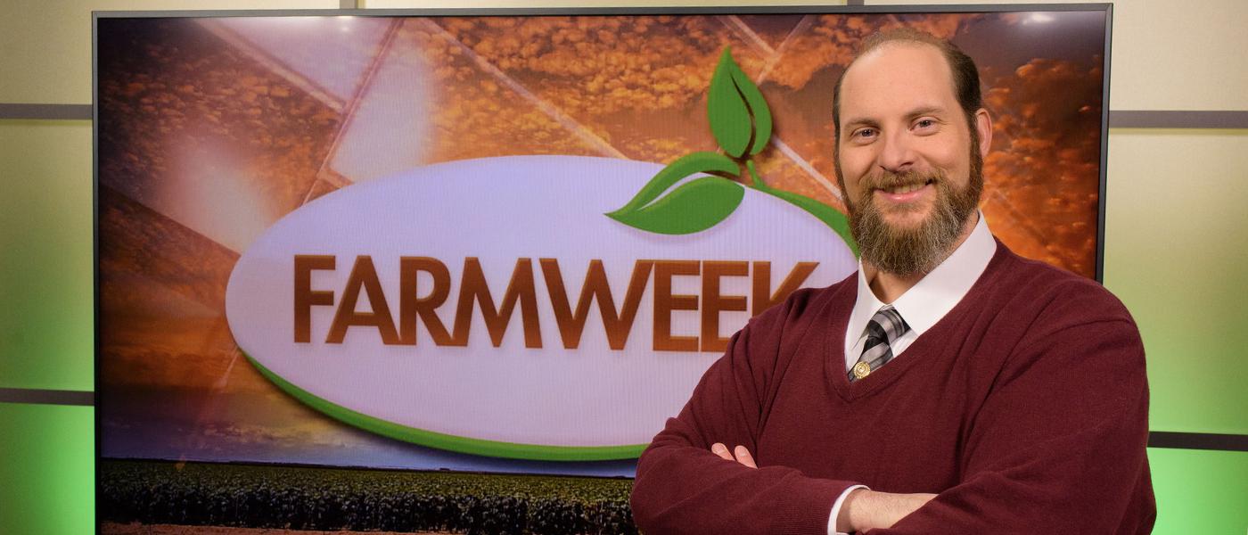 A man stands in front of a TV monitor displaying the Farmweek logo.