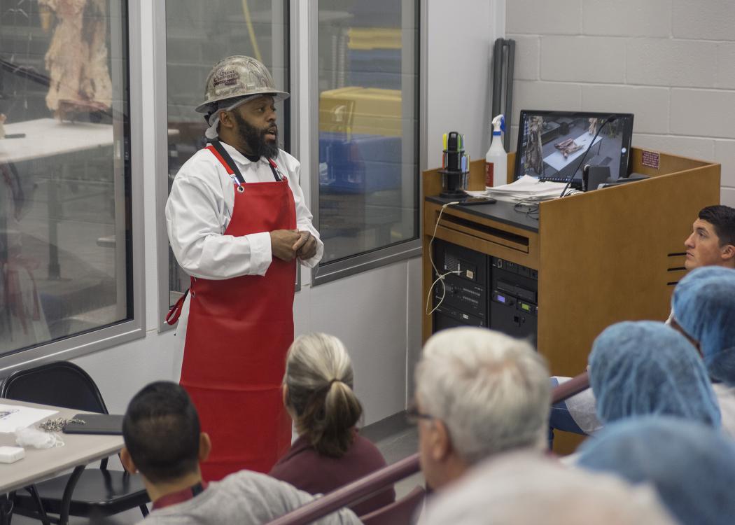 A man in a red apron speaks to a class in a lab.