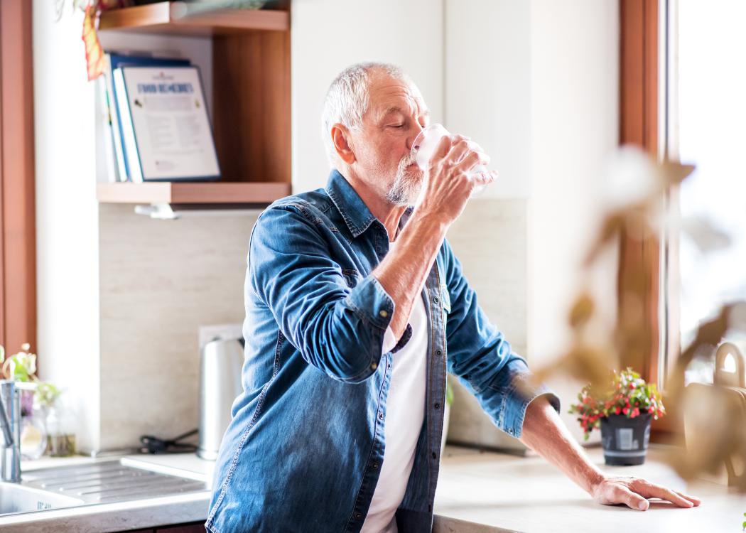 An older man drinks water in the kitchen.