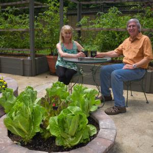 A man wearing an orange shirt and a woman wearing a blue tank top both sitting at a garden table on a concrete slab with planters full of different flowers and plants