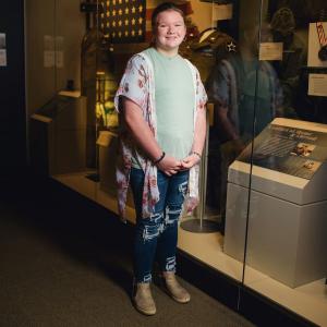 A smiling young girl wearing a floral blouse standing in front of a military museum exhibit.