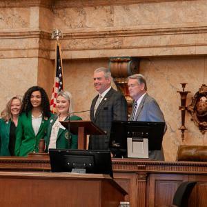 Three women, wearing green blazers, smile for a group photo with two men, wearing suits.