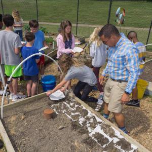 Children lined up and holding paper towels with seeds beside a raised bed garden with paper towels covered in dirt.