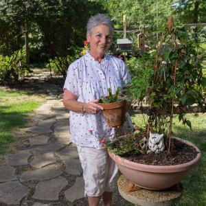 A woman wearing a white shirt with red and blue stars holds a potted plant in a garden.