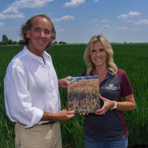 A man and woman stand in a green field holding a book titled “Between the Levees.”