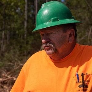 A man wearing a hardhat and orange shirt in front of trees.