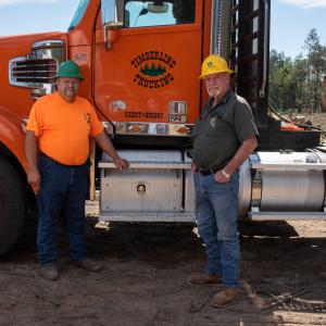Two men wearing hard hats standing in front of an orange logging truck.