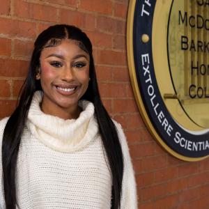 A Black woman wearing a white turtleneck sweater stands smiling beside the University of Mississippi Honor College seal.