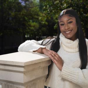 A Black woman wearing a white turtleneck sweater smiling and leaning on a pillar.