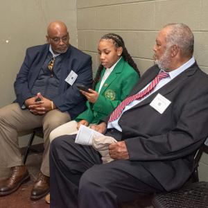 A teen dressed in a green blazer shows two men, on either side, her smartphone.