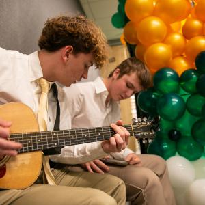 Seated near gold, green, and white balloons, one boy plays guitar while the other sings.