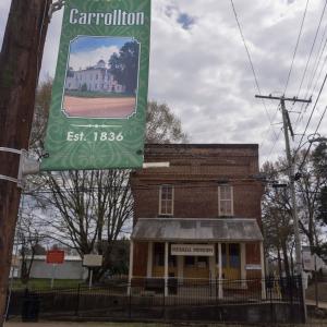A sign that lists “Welcome to Carrollton Est. 1836) in the foreground and the brick Merrill Museum in the background.