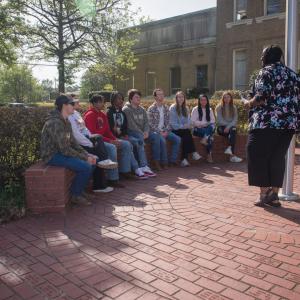 Teens seated outside listening to a woman presenting.