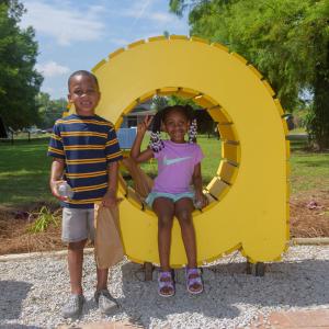 A girl sitting in a yellow lowercase letter “A” and a boy standing beside her.