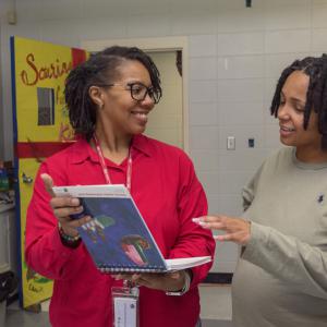 A woman holds a book that she shows to another woman in a classroom.