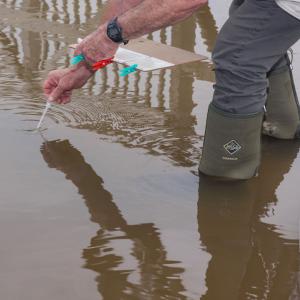 One hand holding a clipboard and another pipetting water out of a waterway.