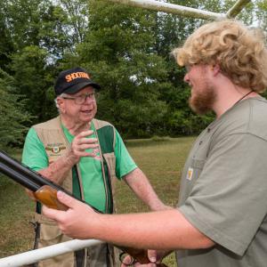 A man wearing a vest and green shirt, talking to a young man smiling and holding a shotgun.