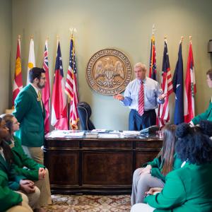 A man in a blue polo shirt and red tie, flanked by the State Seal of Mississippi and national flags, stands behind a desk and talks to seven teens, each wearing green sport jackets.