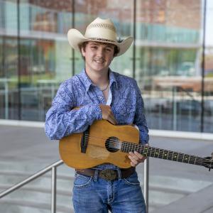 A young man wearing a cowboy hat, paisley collared shirt, and a belt buckle on jeans, holding a guitar and smiling.