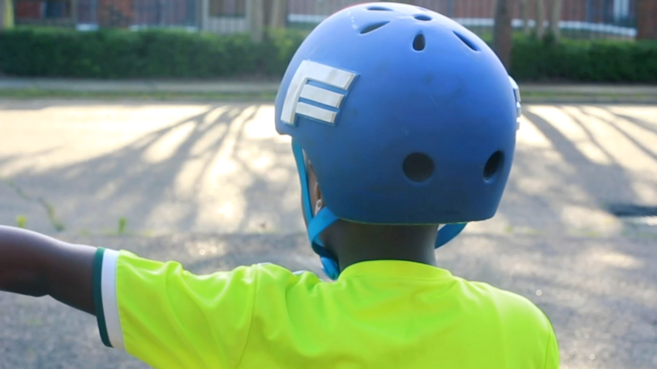 An African-American child wearing a blue helmet and neon yellow shirt extends his left hand to demonstrate a hand signal while riding a bicycle.