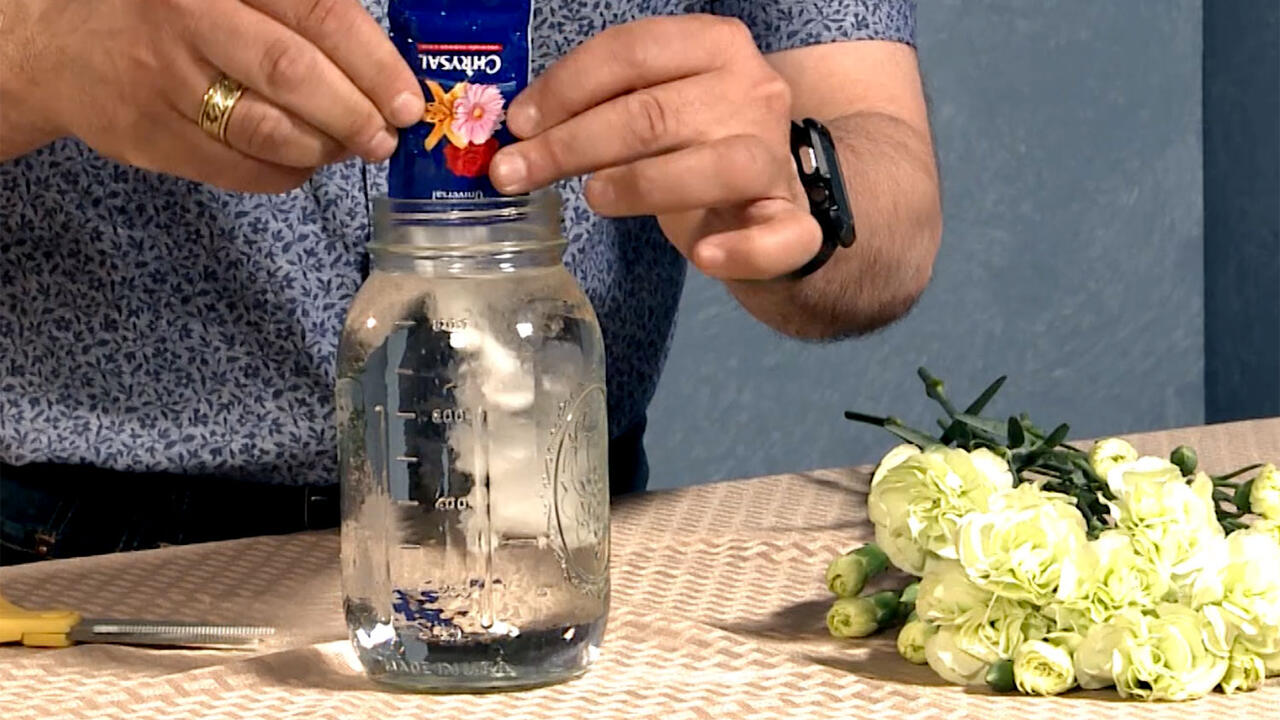 A man in a blue shirt adds a packet of flower food to a clear glass quart jar. White flowers lay beside the jar on a cloth-covered table.