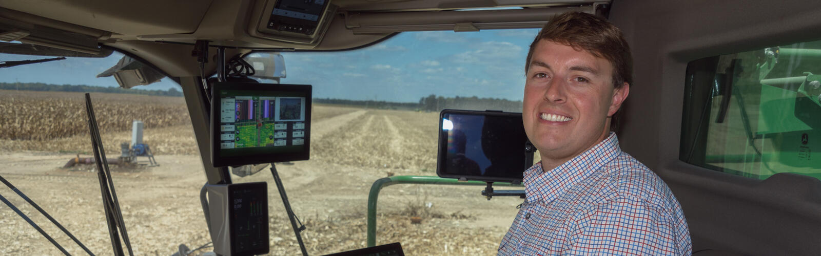 A smiling person seated in a combine in a corn field.