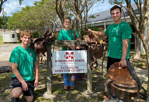 Two boys and a woman, all wearing green shirts, standing with saddles near a Mississippi State University Extension Service sign.
