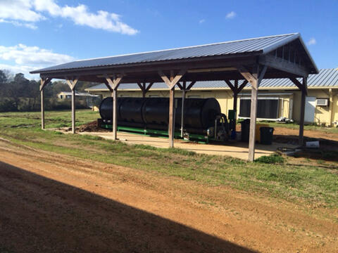 An in-vessel or rotary drum composter underneath a metal roof. The composter is a large metal cyclinder resting on a green metal stand.