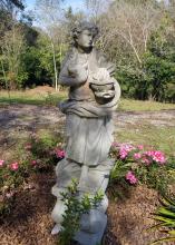 Stone statue of a female holding a container stands in a flower bed.