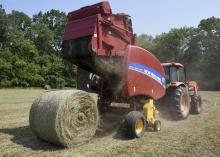 A red baler hitched to the back of an orange tractor drops a new, round bale of hay into a field.