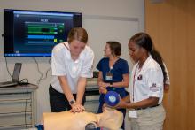 One teenager clasps both of her hands to press on a training manikin’s chest, while another female student holds a ventilator mask over its face. A nurse in the background watches a large wall-mounted monitor.