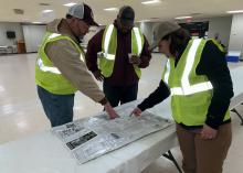 Two men and a woman, all wearing baseball caps and reflective yellow vests, look at a roadmap spread out on a table in a large, empty, well-lit room.
